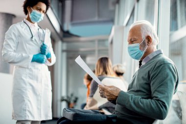 Senior man wearing protective face mask going through his medical data wile sitting in a hallway at the hospital.