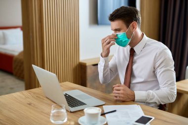 Businessman adjusting protective face mask while working on a computer in hotel room. 