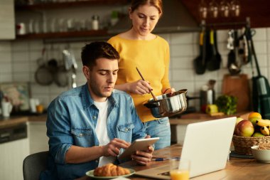 Freelance worker using touchpad and computer while being at home with his wife who is preparing food. 