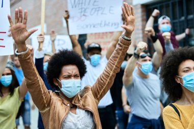 African American woman with raised arms wearing protective face mask while participating in public demonstrations. 