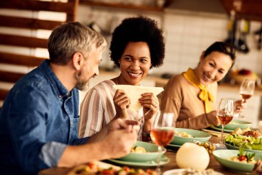 Happy African American woman communicating with her friends while eating lunch in dining room at home.