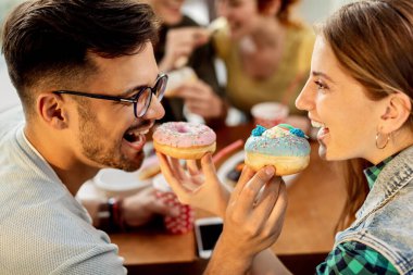 Young couple having fun while sharing their donuts and feeding each other in a cafe. 
