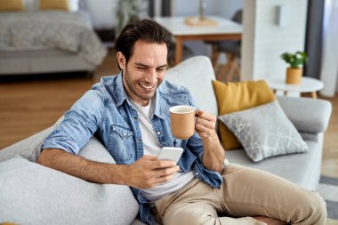 Young happy man texting on smart phone while relaxing on the sofa and drinking coffee. 