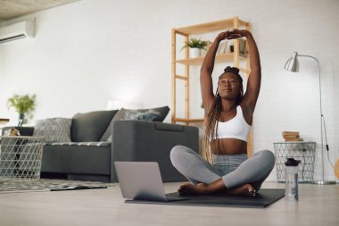 Afrika kökenli Amerikalı sporcu kadın, yoga yaparken dizüstü bilgisayar kullanıyor ve oturma odasında esniyor..