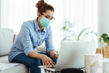 Female freelance worker wearing face mask while using laptop and working from home during virus epidemic. 