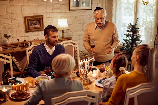 Jewish multi-generation family having a meal at dining table on Hanukkah. Focus is on senior man proposing a toast.