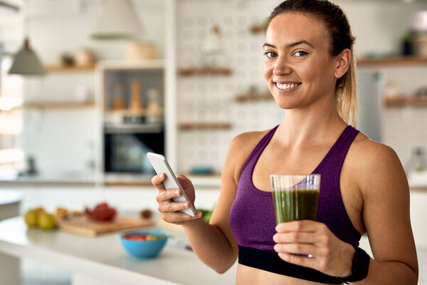 Young happy sportswoman drinking detox smoothie while text messaging on mobile phone in the kitchen. 