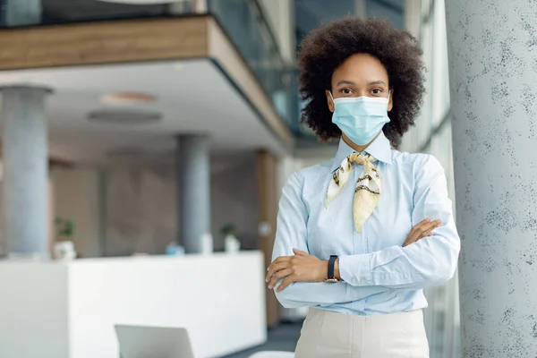 Successful black businesswoman wearing face mask while standing with arms crossed  in the office.