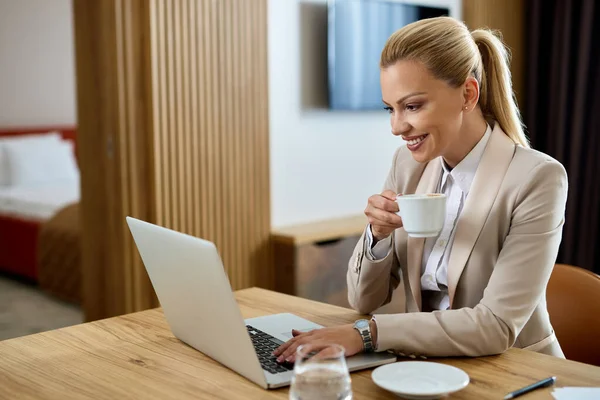 Happy businesswoman surfing the net on laptop while drinking coffee in hotel room.