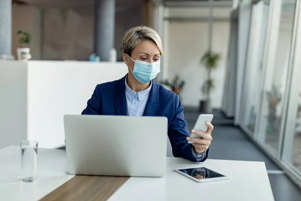 Female entrepreneur working on a computer and texting on cell phone while wearing protective face mask in the office.