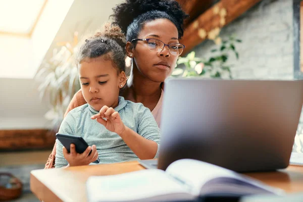 African American stay at home mother working on laptop while daughter is sitting on her lap and using mobile phone at home.