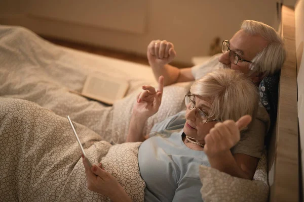 High angle view of mature couple relaxing in bed and surfing the net on digital tablet in the evening,