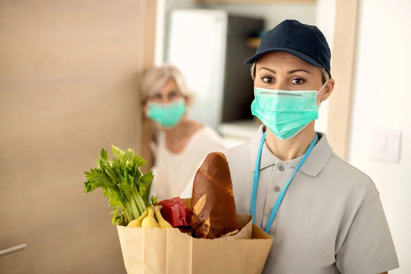 Delivery woman wearing protective face mask while bringing groceries to a senior woman during coronavirus pandemic.