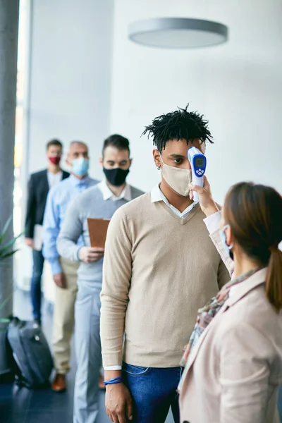Young black man with face mask waiting in line and getting his temperature measured due to coronavirus pandemic.