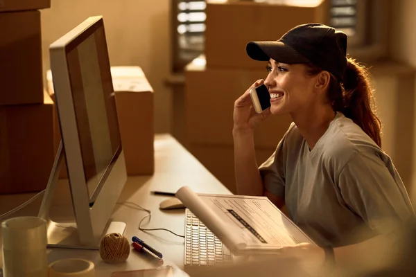 Happy delivery woman reading an e-mail on desktop PC while communicating over cell phone in the office.