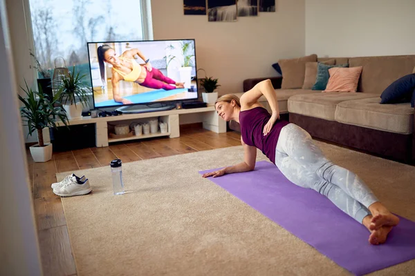 Dedicate athletic woman exercising in side plank pose while watching fitness class on a TV at home.