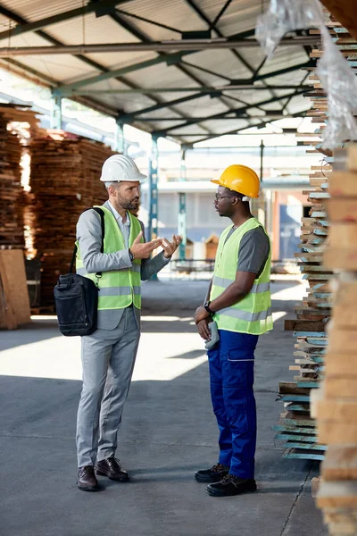 African American warehouse worker communicating with inspector and getting instructions from him at timber distribution department.