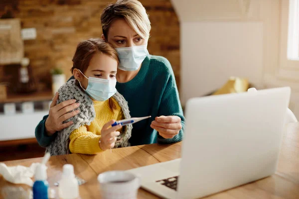 Single mother with face mask having video call with a pediatrician while her daughter is having a fever at home.