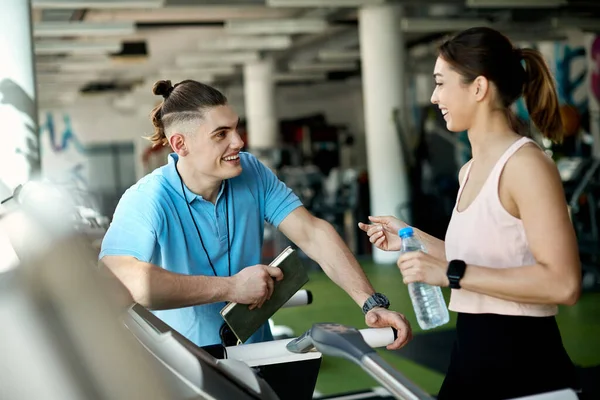 Happy personal training communicating with athletic woman who is walking on treadmill in a gym.