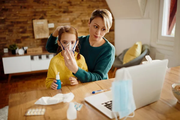 Caring mother using nebulizer during inhaling therapy of her ill daughter while following instructions on a computer at home.
