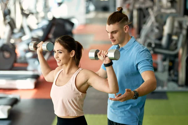 Young athletic woman exercising with hand weights with help of personal trainer in a gym.