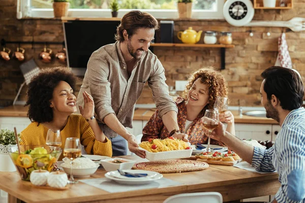 Young happy man serving food at dining table while having lunch with friends at home. 
