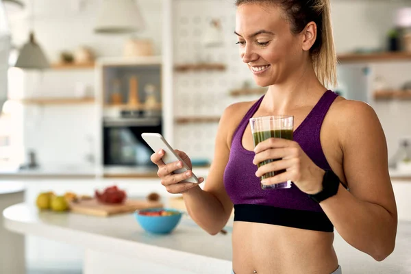 Young happy sportswoman using smart phone while drinking detox smoothie in the kitchen.