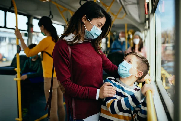 Happy mother communicating with her son while commuting by public transport and wearing face masks due to COVID-19 pandemic.