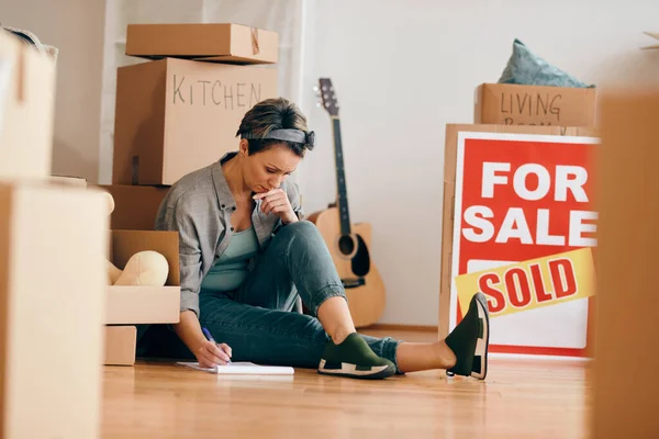 Displeased woman sitting on the floor among cardboard boxes and filling paperwork after selling her home.