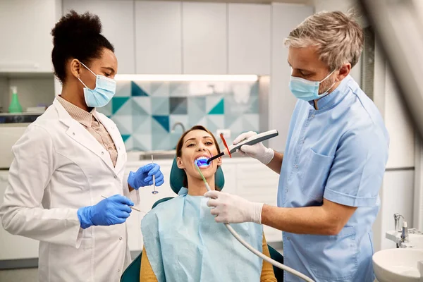 African American dentist and her assistant using ultraviolet lamp during dental procedure of female patient.