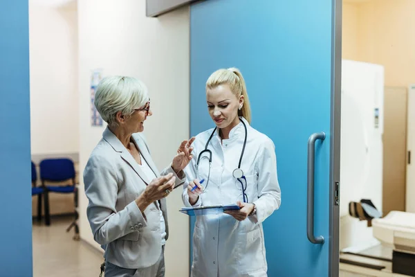 Happy doctor reading medical reports of her senior patient while talking to her in a lobby at the hospital.