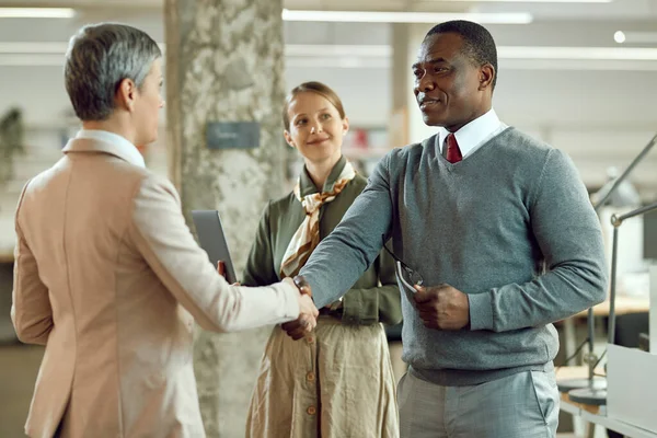 Happy African American corporate manager shaking hands with businesswoman while greeting her in the office.
