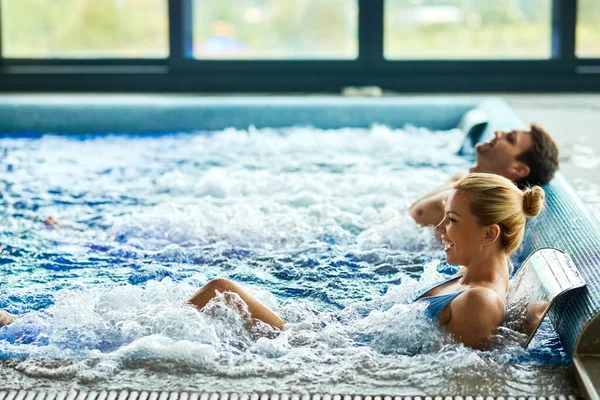 Happy woman having fun while enjoying with her husband in water massage in the pool at wellness center. Copy space. 