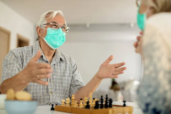 Happy mature man with face mask talking to his wife while playing chess at home during COVID-19 epidemic.