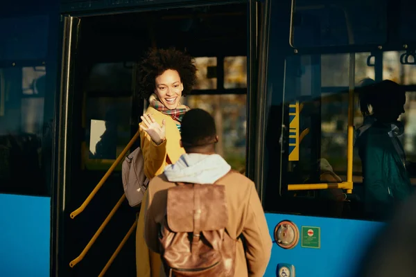 Happy black woman entering a bus and waving to her boyfriend on the station.