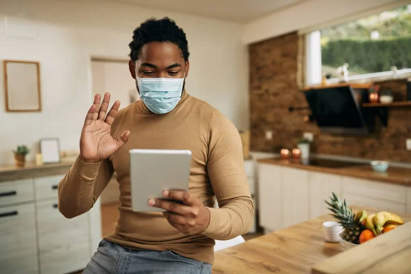 African American man with face mask using digital tablet and greeting someone during a video call from home.