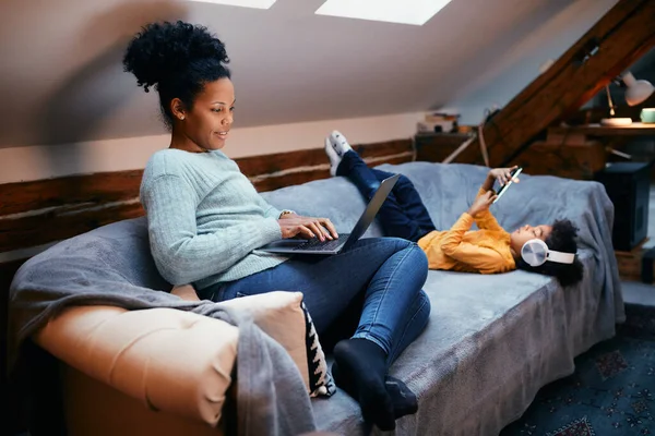 Happy African American working mother using laptop on the sofa while her daughter is listening music over mobile phone.