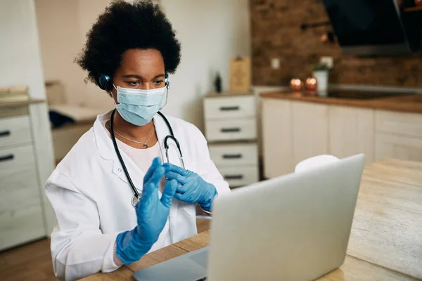 Black female doctor wearing face mask while using laptop and talking during conference call from her office.