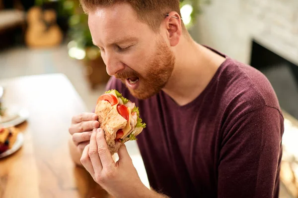 Young man tasting sandwich with eyes closed while eating at home.