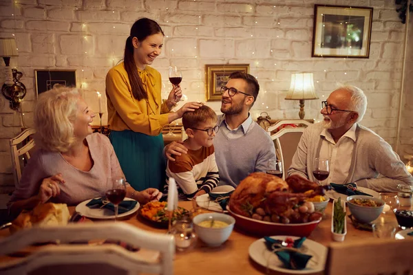 Cheerful woman proposing Thanksgiving toast while celebrating with her extended family during a meal at dining table.