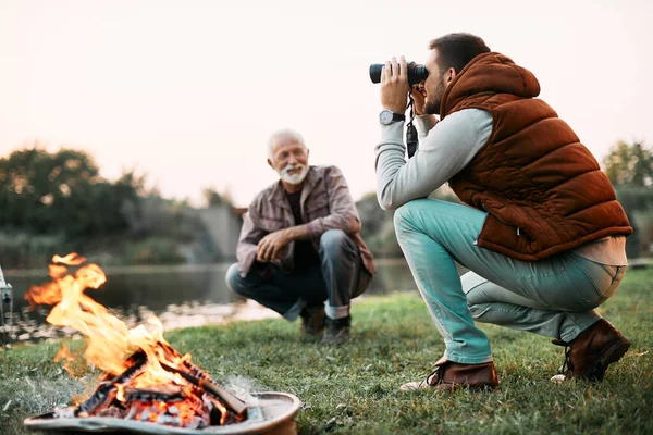 Adult man camping with his senior father by the river and looking through binoculars. Copy space.