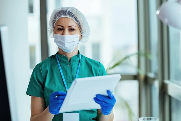 Female doctor wearing face mask and gloves while analyzing medical data at clinic and looking at camera.
