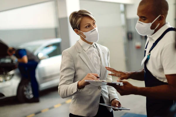 Businesswoman and African American repairman wearing protective face masks while talking and going through reports in car service workshop.