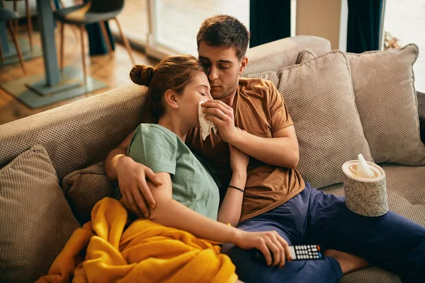 Young woman crying while watching sad movie on TV with her boyfriend who is wiping tears from her face.