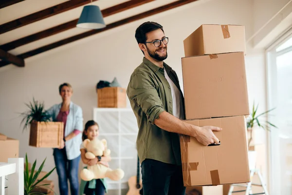 Happy father carrying stack of cardboard boxes while relocating in a new apartment. His wife and daughter are in the background. 