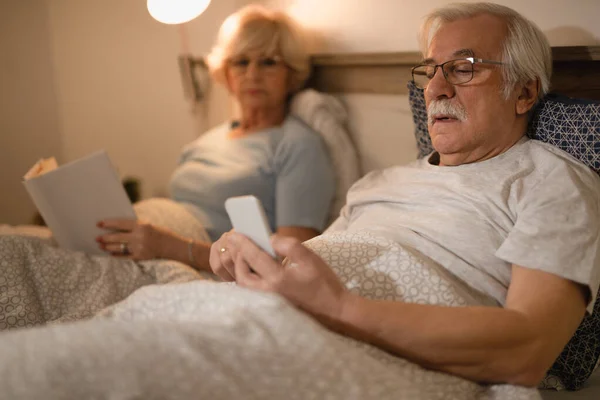 Mature man text messaging on mobile phone while resting in bed with his wife in the evening.