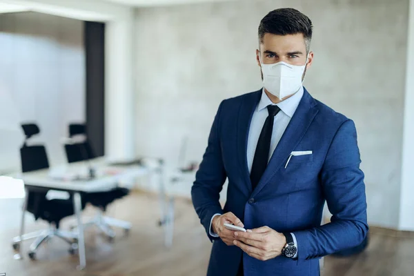 Smiling businessman wearing protective mask on his face while using mobile phone in the office and looking at camera.