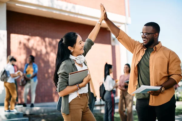 Happy Asian student giving high-five to her black male friend while meeting at the university.