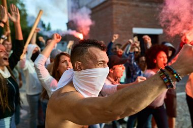 Furious crowd of people raising a revolution against government on city streets. Focus is on shirtless man. 