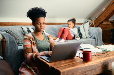 African American mother using laptop and working at home while her daughter is surfing the net on touchpad on the sofa.